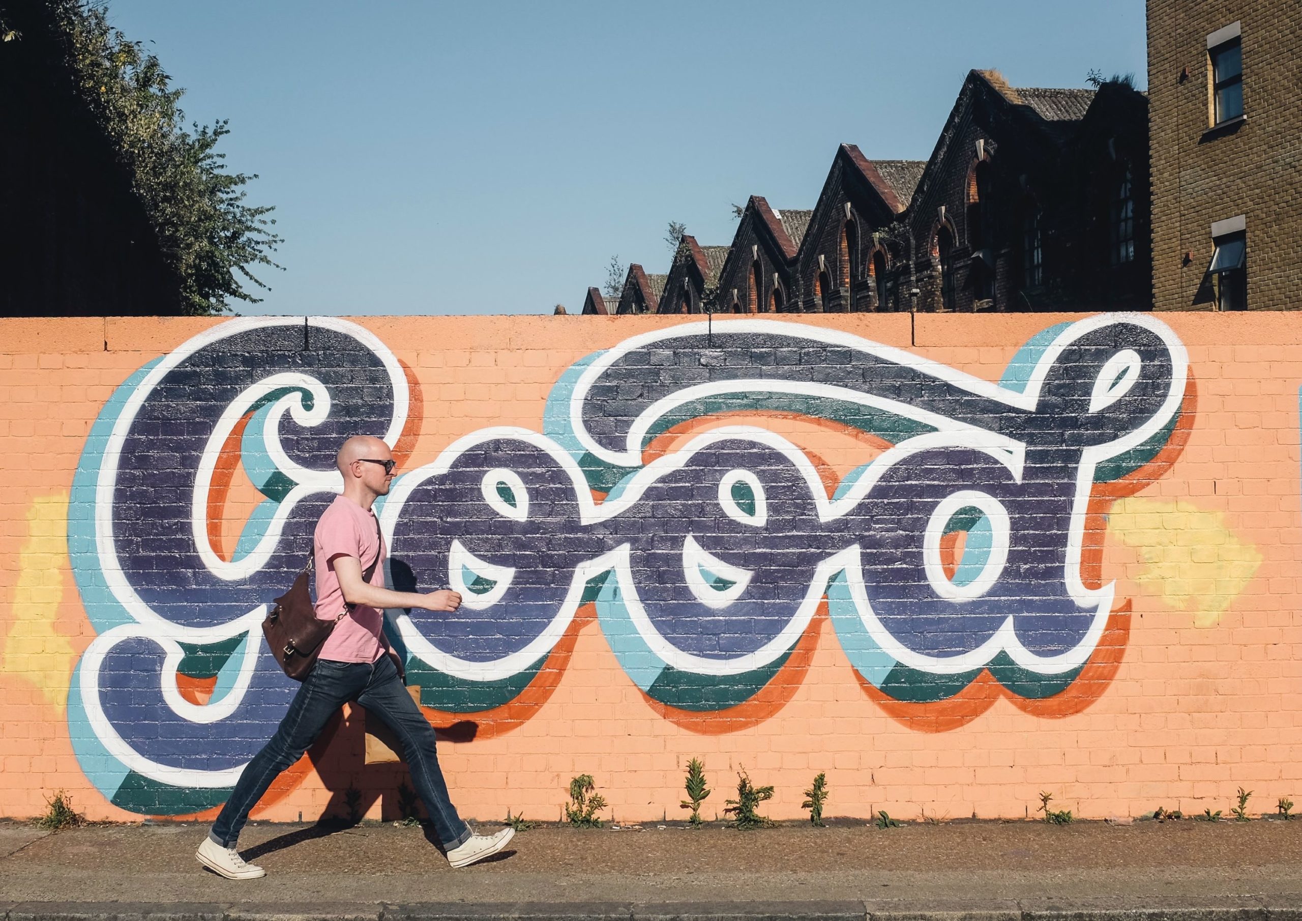 A man walks along a sunny city street with colourful graffiti artwork in the background that reads, "Good".